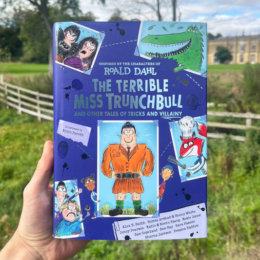 A hand holds a book titled 'The Terrible Miss Trunchbull and Other Tales of Tricks and Villainy' by Roald Dahl. The book cover is predominantly blue with various colorful cartoon illustrations and text.
