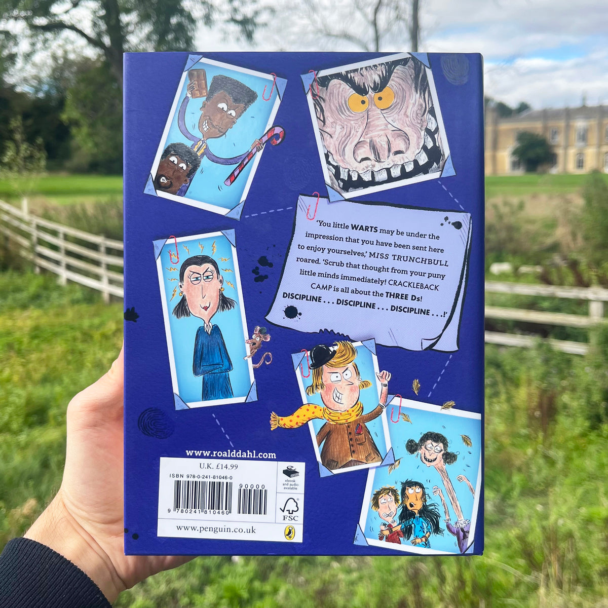 A hand holds a blue book cover in an outdoor grassy field. The cover features colorful cartoon illustrations and a white text box with a barcode and website details.
